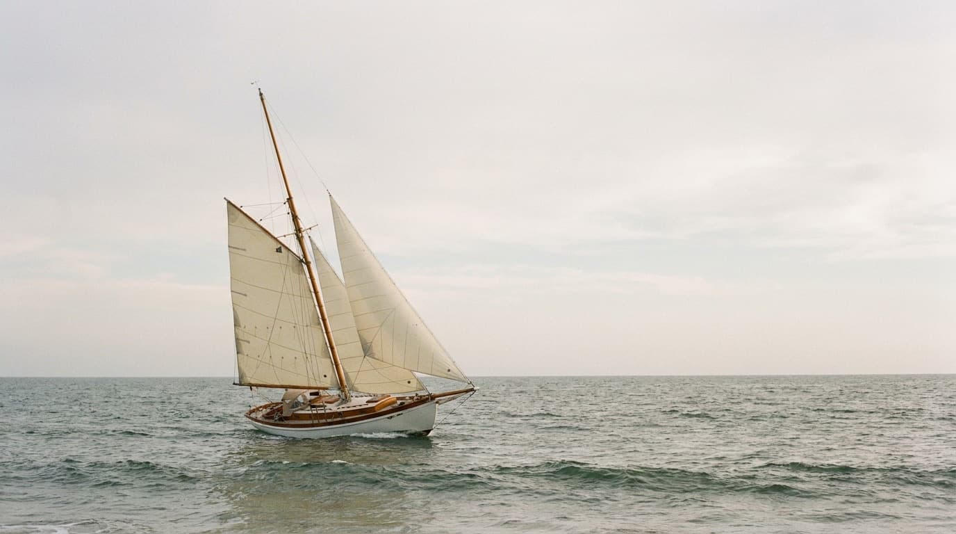 A heavy full-keel double-ended cruising sailboat of about 32 feet under working sail in open water, heeled gently, carrying a mainsail and two headsails set from a long bowsprit in a cutter rig, with salt-worn white topsides and varnished timber trim under soft overcast light.